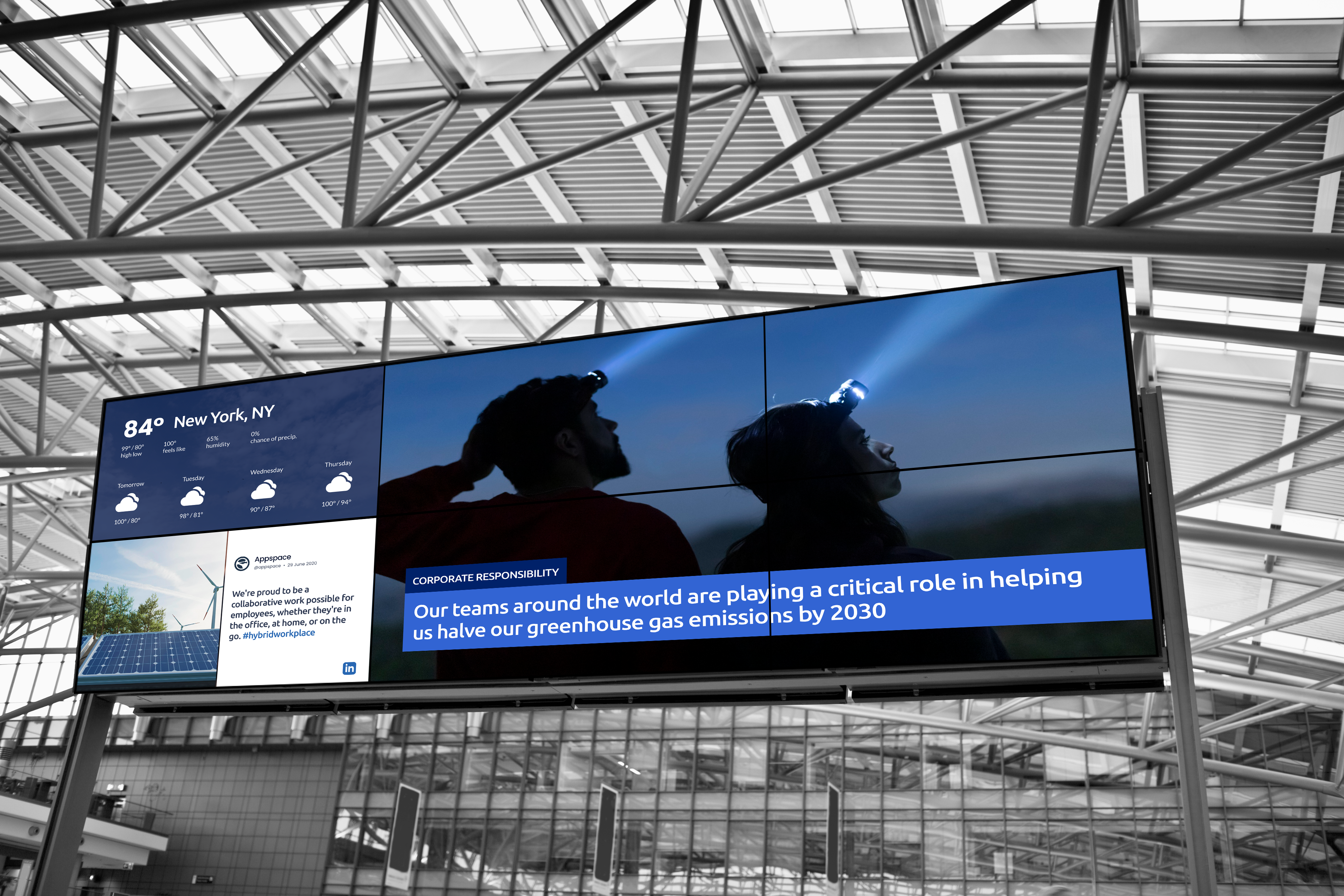 A large digital display in an airy, modern building shows weather for New York, NY, news highlights, and a photo of two people wearing headlamps. Text below discusses reducing greenhouse gas emissions by 2030.