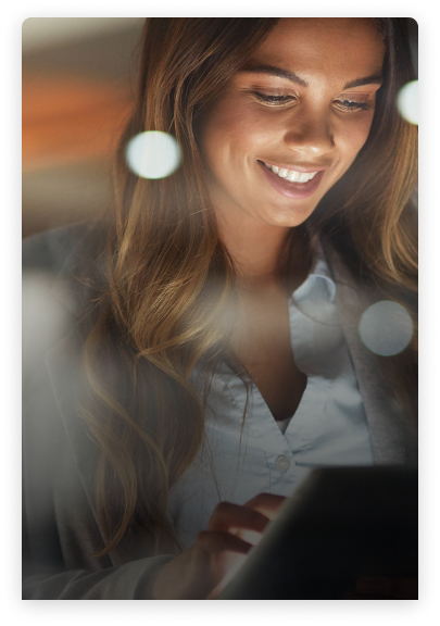 A woman with long brown hair is smiling while looking at a tablet. She is wearing a white shirt and a gray blazer. The background is softly blurred with bokeh lights.