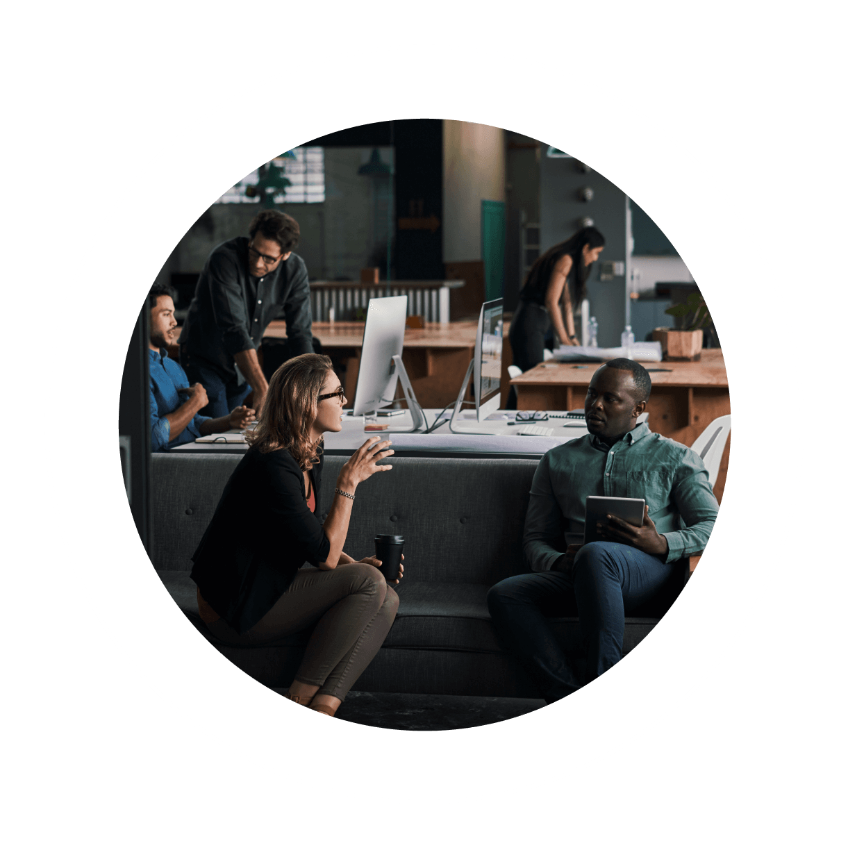 A group of people work in an open office space. Two are seated on a couch having a conversation; one is holding a tablet. In the background, two more people work at desks with computers. The environment is modern and collaborative.