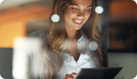 A woman with long hair smiles while using a tablet. She is indoors, and the photo is taken through a glass surface, creating soft reflections and light spots.