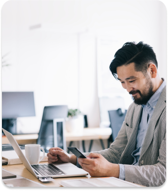 A person with short dark hair and a beard is sitting at a desk in an office. They are holding a smartphone in one hand and have a laptop open in front of them. A white mug is also on the desk. The setting is bright and modern.
