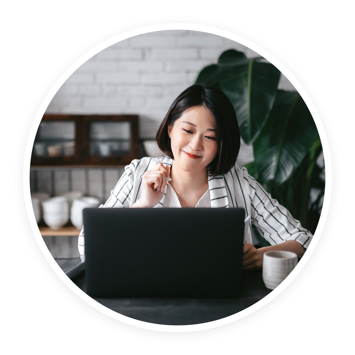 A woman in a striped blazer is sitting at a desk, smiling while looking at a laptop. She holds a pencil in one hand, with a coffee cup nearby. The background features a white brick wall and large green plant leaves.